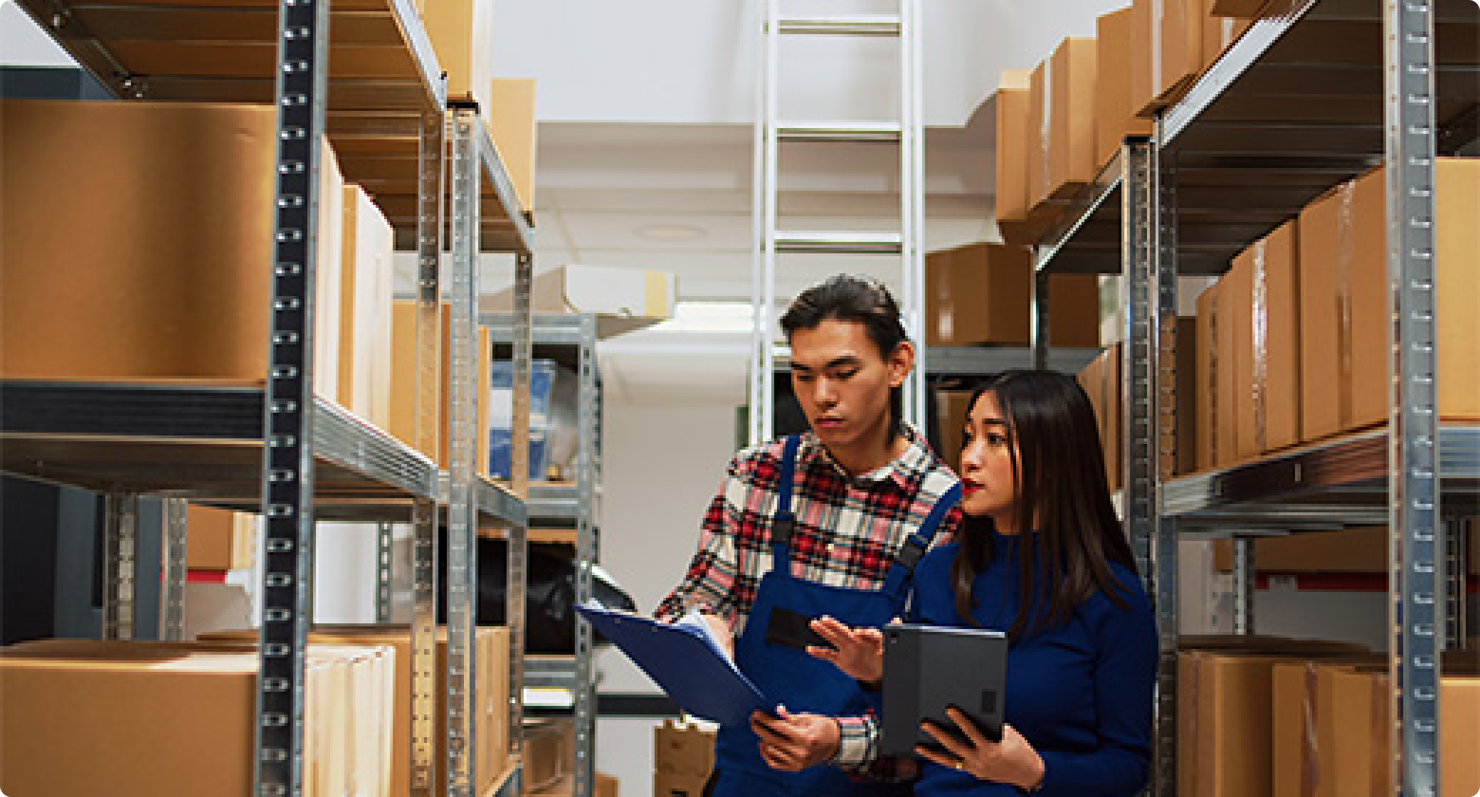Two warehouse staff standing among shelves of boxes, reviewing inventory details using a clipboard and tablet during a discussion about stock.