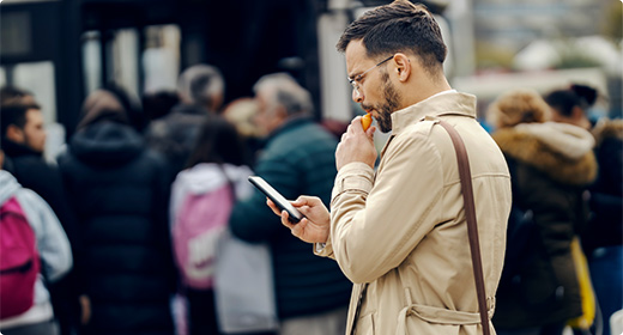 Man in beige coat standing outdoors checking smartphone with thoughtful expression while crowd of people move in the background.